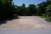 Photo: Buttonwood: Large, gravel, partially shaded campsite with picnic table and grill, bordered by a wood fence and surrounded by bushes.