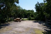 Photo: Buttonwood: Small, gravel, partially shaded campsite with picnic table and grill, bordered by a wood fence and surrounded by bushes.