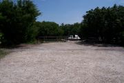 Photo: Buttonwood: Gravel, partially shaded campsite with picnic table and grill, bordered by a wood fence and surrounded by bushes. Boat at dock in the background.