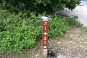 Photo: Buttonwood: Site number post with white numbers and arrows on a brown background, bushes in background.
