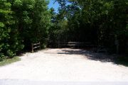 Photo: Buttonwood: Partially shaded gravel campsite with picnic table and grill, bordered by a wood fence and surrounded by bushes.