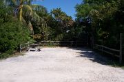 Photo: Buttonwood: Partially shaded gravel campsite with picnic table and grill, surrounded by a wood fence and bushes.