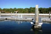 The photograph shows a view of boat slips in the Caladesi Island marina. An aluminum floating dock features power and water pedestals, with an aluminum seawall and green palm trees in the background. Concrete pilings support the docks.  Finger docks protrude out from each side of the main dock at intervals of every two boat slips.