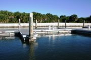 The photograph shows a view of boat slips in the Caladesi Island marina. An aluminum floating dock features power and water pedestals, with an aluminum seawall and green palm trees in the background. Concrete pilings support the docks.  Finger docks protrude out from each side of the main dock at intervals of every two boat slips.
