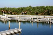 The photograph shows a view of boat slips in the Caladesi Island marina. An aluminum floating dock features power and water pedestals, with an aluminum seawall and green palm trees in the background. Concrete pilings support the docks.  Finger docks protrude out from each side of the main dock at intervals of every two boat slips.