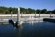 The photograph shows a view of boat slips in the Caladesi Island marina. An aluminum floating dock features power and water pedestals, with an aluminum seawall and green palm trees in the background. Concrete pilings support the docks.  Finger docks protrude out from each side of the main dock at intervals of every two boat slips.
