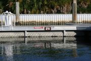 The photograph shows a view of boat slips in the Caladesi Island marina. An aluminum floating dock features power and water pedestals, with an aluminum seawall and green palm trees in the background. Concrete pilings support the docks.  Finger docks protrude out from each side of the main dock at intervals of every two boat slips.