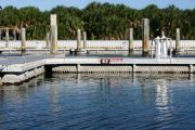 The photograph shows a view of boat slips in the Caladesi Island marina. An aluminum floating dock features power and water pedestals, with an aluminum seawall and green palm trees in the background. Concrete pilings support the docks.  Finger docks protrude out from each side of the main dock at intervals of every two boat slips.