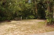 The wooden site marker is at the right. This site has green grass and tan sandy soil. Itâs shaded and surrounded by green vegetation. The brown metal ground grill is to the left, a wooden picnic table and gray electrical box center, and the water spigot to the right.