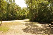 The view from this site shows a paved front road and surrounded heavily with wooded with green vegetation for optimum privacy. The ground grill and the picnic table are also visible to the right of the picture. The site marker is to the left.