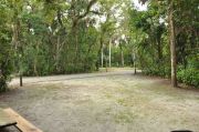 This view from the back of the site shows a paved front road, wooden site marker to the left, sandy soil with sparse green grass and a view into the opposing camp site. A tall palm tree is at the far right side.