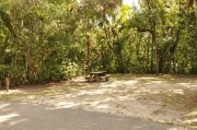 The wooden site marker is to the far left. This site has sparse green grass and tan sandy soil. Itâs shaded and surrounded by green vegetation. The gray electrical box is far right. The wooden picnic table is near the back with the brown metal ground grill behind it.
