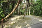 The wooden site marker is at the left. The site has green grass and tan sandy soil, shaded and surrounded by green vegetation. A brown metal ground grill, a wooden picnic table, gray electrical box, and water spigot are right of center.