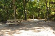This view shows a brown metal ground grill and wooden picnic table to the left. A gray electrical box is near the back center. A water spigot is to the right. It is partially shaded with green vegetation, Oak trees and Cabbage Palms. Soil is tan with sparse green grass.