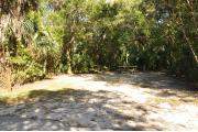 The wooden site marker is at the left. This site has green grass and tan sandy soil. Itâs shaded and surrounded by green vegetation. The brown metal ground grill is to the left, a wooden picnic table and gray electrical box center, and the water spigot to the right.