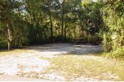 The wooden site marker is at the left. This site has green grass and tan sandy soil. Itâs shaded and surrounded by green vegetation. The brown metal ground grill is to the right, a wooden picnic is center. The front road is paved.