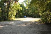 This view from the back of the site shows a paved front road, wooden site marker to the right, sandy soil with sparse green grass and partial view into an opposing camp site with a small blue tent. Cabbage Palms and green vegetation shade this site.