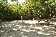 This is a view of the brown ground grill, gray electrical box, water spigot and wooden picnic table. The soil is sandy with sparse green grass. Green vegetation surround the site and gives partial shade.