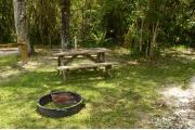 This view from the back of the site shows a paved front road, wooden site marker to the left, sandy soil with sparse green grass and view into opposing camp sites. The wooden picnic table and ground grill is to the right. The information kiosk is across the road center.