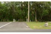 The wooden site marker is at the left. This site is paved and has green grass and tan sandy soil. Two trees line the driveway. Wooden picnic table, and stand up grill are to the right. Gray electrical box and water spigot are near the back left. This is an ADA site.