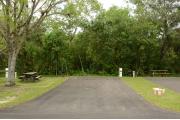 This photo shows the paved driveway with a wooden picnic table, gray electrical box, water spigot, a tree, and green vegetation towards the back of the site. This site has green grass and some tan soil.