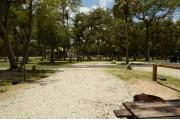 This view shows the brown ground grill, water spigot, gray electrical box and wooden picnic table. The soil is gray to tan color. Green palms are in the background.