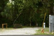 The site is open with limited shade. The view shows the site marker, wooden picnic table, brown ground gill, gray electrical box, water spigot, electrical pole, and guide wires to the right. Green grass, gray/tan soil and green vegetation are at the back of the site.