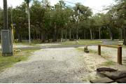 This view shows a wooden picnic table, brown ground grill, water spigot and gray electrical box to the left. The soil is gray to tan color. Green palms are in the background. A guide wire is to the left.