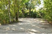 The wooden site marker is to the left. This site has sparse green grass and tan sandy soil. Itâs shaded and surrounded by green vegetation. The brown metal ground grill, a wooden picnic table, gray electrical box and the water spigot are back center.