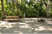 This is a view of a wooden picnic table, gray electrical box, water spigot and a brown ground grill. The soil is sandy with sparse green grass. Green palms and Oak trees surround the site.