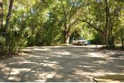 This view from the back of the site shows a paved front road, wooden site marker to the right, sandy soil with sparse green grass, and view into an opposing camp site with a white truck and blue tent. A wooden picnic table is to the right. Site #015 adjoins to the left.