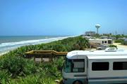There is substantial scrub located on the face of the dune. A wooden observation deck is located on top of the dune. The beach is off to the left with blue skies. A motor home is parked right beside the observation deck.