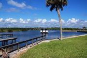 A boat launch and basin located on the Intracoastal Waterway. There is green grass, a palm tree and blue skies with beautiful cumulus clouds. A catwalk runs along both sides of the boat launch.