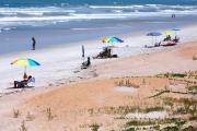 Blue skies and ocean waves at low tide. Natural dunes and coquina sand comprise the beach. Beachgoers with chairs and umbrellas enjoy the day.