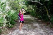 A person is looking through binoculars at wildlife with scrub oaks and saw palmetto bushes around them. The Â¼ mile nature trail goes through the woods exposing wildlife and native plants.