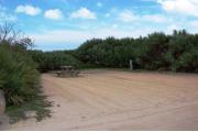 Packed sand campsite with low palmetto plants and sea oats along the back. Picnic table set off to the left side with grill behind it. Electric and water hookup located to the right. Palmetto plants border the right side for privacy.