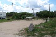 Packed sand campsite with no privacy vegetation between sites. Picnic table and ground grill located on the left. Barricade posts in back of site. Palmetto plants provide a thick buffer to the back. Electric and water hookups located to the right.