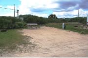 Packed sand campsite with no privacy vegetation between sites. Picnic table and ground grill located on the left. Barricade posts in back of site. Palmetto plants provide a thick buffer to the back. Electric and water hookups located to the right.