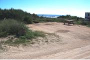Packed sand campsite with low palmetto plants and sea oats along the back.  The ocean is visible. Low vegetation borders the right side. Electric and water hookups located to the right. Picnic table is on the right of the site and ground grill is located at the back of site.