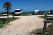 This picture is taken from the back looking out towards campground road.  Packed sand campsite with no privacy vegetation between sites. Picnic table and ground grill located on the left. Barricade posts in back of site. View to the ocean.  Electric and water hookups located to the right.