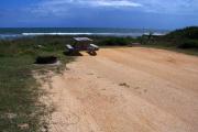 Packed sand campsite with low scrub and sea oats lining the back of the site. Ocean is visible. Picnic table and ground grill off to the left. Low vegetation along both sides of site.