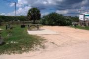 Packed sand campsite with large palmetto plants with palm tree. Low vegetation between sites. Handicap-accessible picnic table sits on a concrete pad and accessible ground grill off to the left of the campsite. Electric and water hookups on right side. Palmetto plants provide a buffer to the back.