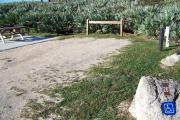 Packed sand campsite with palmetto plants along the back providing a buffer.  Handicap-accessible picnic table sits on a concrete pad. Accessible ground grill off to the left. Barricade posts to protect vegetation.