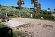 Packed sand campsite with palmetto plants along the back providing a buffer.  Handicap-accessible picnic table sits on a concrete pad. Barricade posts to protect vegetation. Electric and water hookups on right.