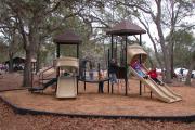 View shows multiple children playing on playground. Some children are sliding down slides, of which there are two. There are also two towers with a foot bridge connecting them. The area around the playground has a plastic border and the enclosed area is covered with mulch and shaded by trees.