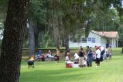 View shows people of various ethnic backgrounds picnicking on a grassy slope. There are people grilling and eating at picnic tables. The lawn is partially shaded by tree cover, and the Recreation Hall facility can be seen in the background.