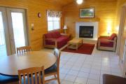 View of the living room and dining room of a modern cabin. There are two couches and a coffee table in the living room nestled in front of a fire place. The walls are wood and the floors are tile.