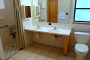 View of the bathroom in a modern cabin. There is an ADA compliant toilet, a dual sink vanity style counter with a large low mirror, and a walk-in/roll-in shower equipped with grab bars and a fold down chair. The walls are white and the floors are tile.