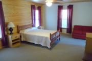 View of the bedroom in a modern cabin. There is a full size bed, a dresser, and a hide away couch that sleeps 1 person. The floors have short pile carpet and the walls are a combination of wood and white paint.