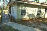 View of the back of a modern ADA compliant cabin. A concrete sidewalk leads to a wood ramp that enters the cabin through a large screen porch. The cabin is grey with white trim and is wood frame constructed.